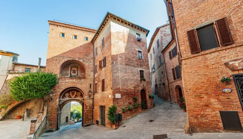 Antica porta in mattoni a Torrita di Siena, con un affresco e un vicolo acciottolato che sale tra case medievali.