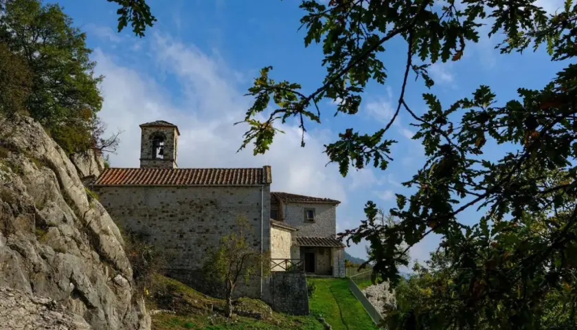 L'eremo di Cerbaiolo, con la sua pietra antica e il campanile, si staglia contro un cielo azzurro tra i rami di quercia.