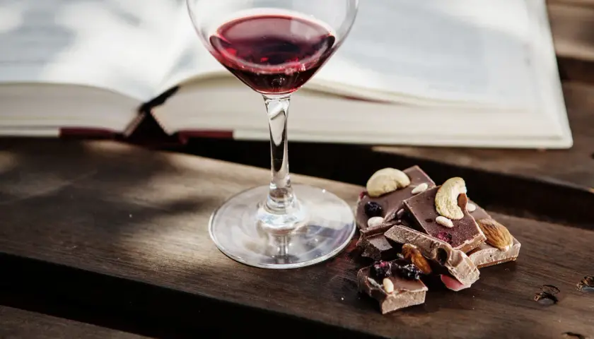 Un bicchiere di vino rosso accanto a pezzi di cioccolato con frutta secca e noci. Un perfetto abbinamento vino e cioccolato per una serata di relax.