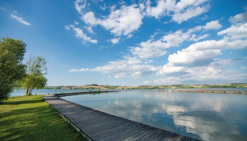 Passerella in legno su un lago tranquillo, con colline ondulate sullo sfondo. Un luogo idilliaco in Valdichiana, dove la natura incontra la serenità.