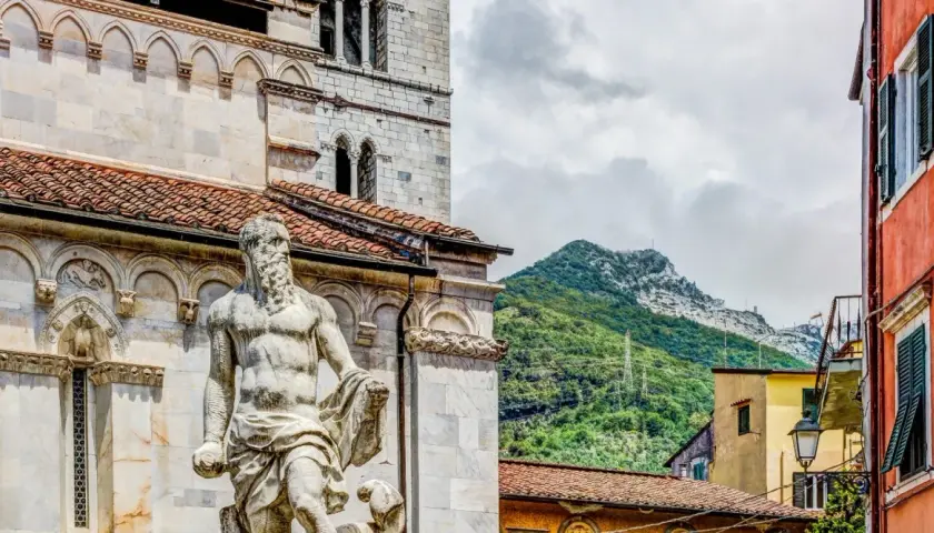 Statua nel carrara centro storico, con montagne verdi sullo sfondo e case colorate.