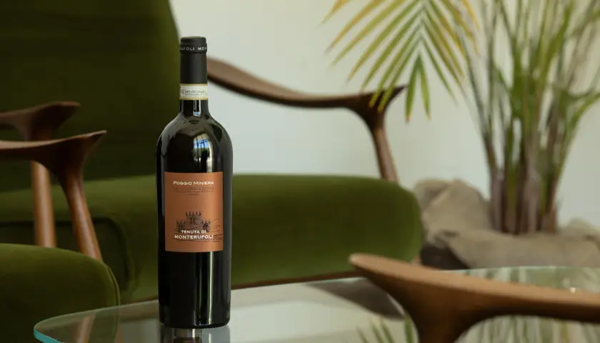 Bottiglia di vino rosso Poggio Miniera, un'eccellenza tra i vini toscani, su un tavolino di vetro, con poltrone verdi e piante sullo sfondo.