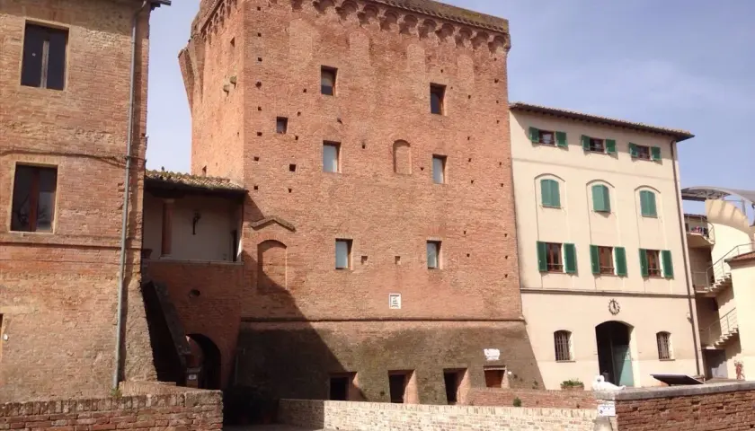 Torre medievale in mattoni a Monteroni d'Arbia, con edifici adiacenti in pietra e intonaco.