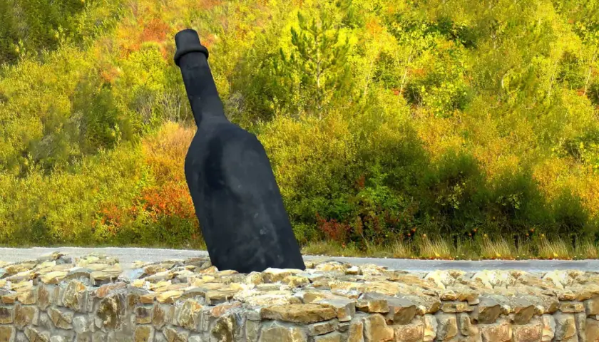 Giant black wine bottle sculpture emerging from a stone wall, with a colorful autumn forest in the background.