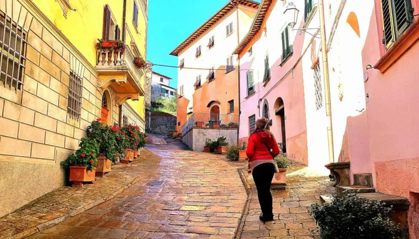 Una donna cammina su una strada acciottolata a Palaia, Pisa, tra case colorate e vasi di fiori rossi.