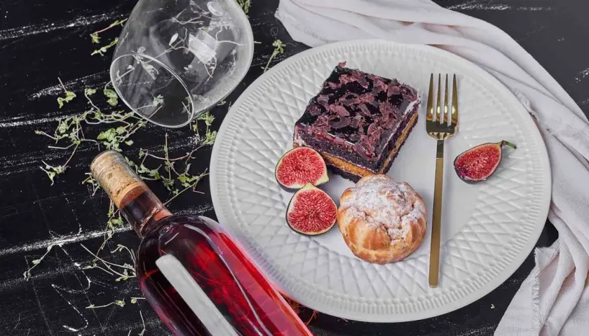 Un bicchiere rovesciato, una bottiglia di vino rosso e un piatto con torta al cioccolato, bignè, fichi e posate dorate, un'invitante composizione per gustare vini da dessert.