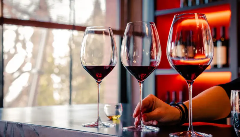 Calice di vino rosso su un bancone, con una mano che sfiora uno stelo. L'atmosfera è quella della cantina il poggio, con luci soffuse e bottiglie sullo sfondo.