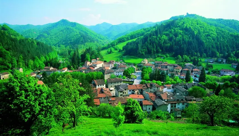 Un borgo incantevole, Palazzuolo sul Senio, immerso nel verde lussureggiante delle colline appenniniche. Case con tetti rossi si aggrappano ai pendii, circondate da alberi rigogliosi e un cielo azzurro.