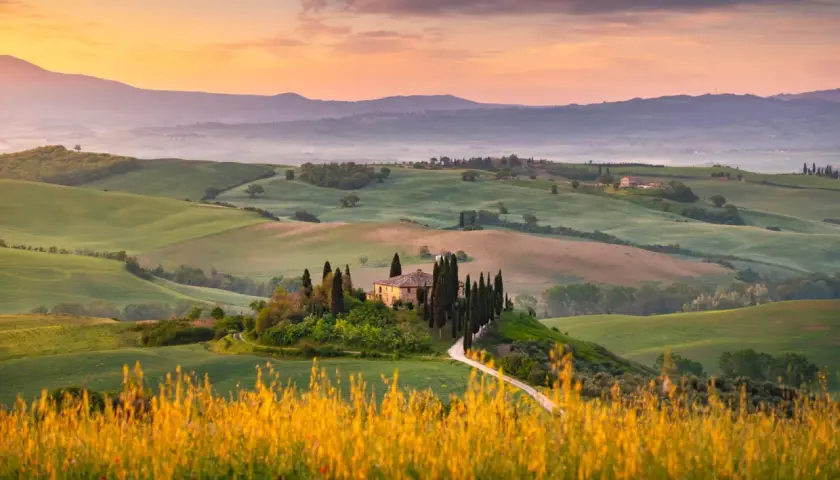 Dolci colline verdi nel Val d'Orcia, con una casa colonica e cipressi su una collina, al tramonto.