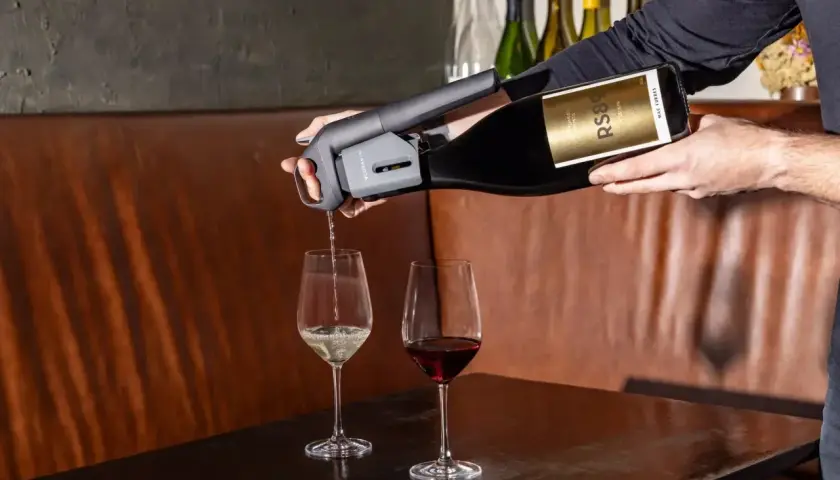 Versando vino bianco da una bottiglia con un erogatore Coravin, accanto a un bicchiere di vino rosso. Un tipo di vino da scoprire.