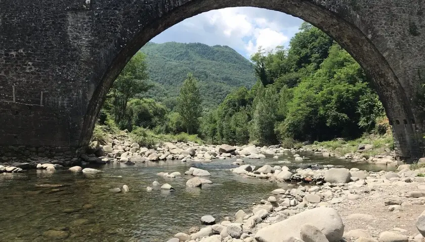 Il ponte di Castruccio, con il suo arco maestoso, sovrasta un fiume serpeggiante tra rocce e vegetazione lussureggiante.