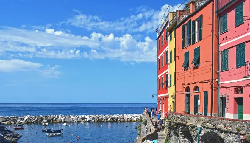 Case, a Firenze c'è il mare! Case colorate affacciate su un porto con barche, sotto un cielo azzurro con nuvole bianche.