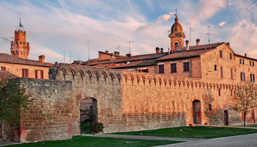 Antica cinta muraria in pietra delle Crete Senesi, con torri medievali e cielo al tramonto.