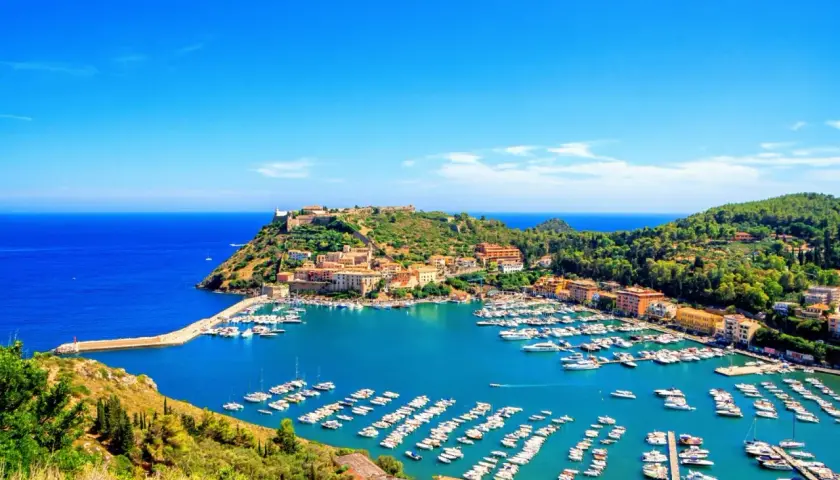 Porto Ercole cosa vedere: un incantevole porto con barche, case colorate e un castello su una collina verde, bagnato da un mare azzurro.