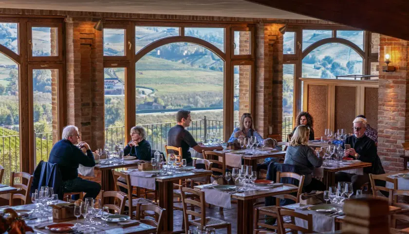 Persone cenano in un ristorante con vista panoramica su un'azienda agricola marrone. Tavoli apparecchiati con cura, mattoni a vista e grandi finestre creano un'atmosfera accogliente.