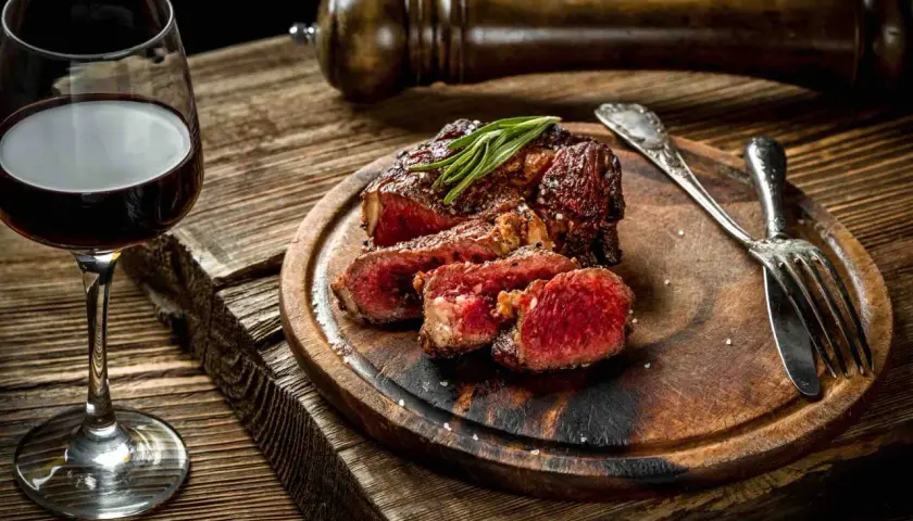 Bistecca succulenta con rosmarino, accompagnata da un bicchiere di vino tannico e posate d'argento su un tagliere di legno rustico.