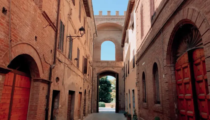 Un vicolo antico con edifici in mattoni rossi e un arco monumentale che conduce a un giardino. Sembra un buon convento.