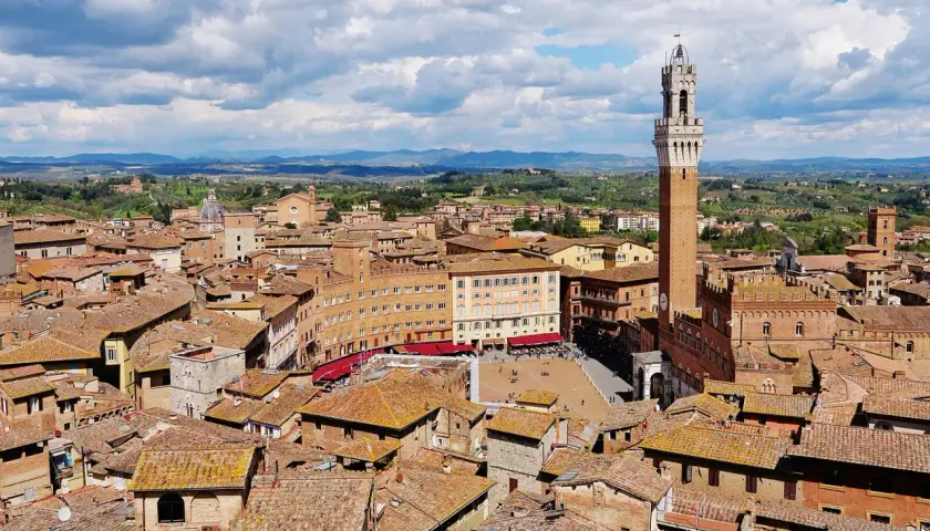 Vista aerea di Siena con la Torre del Mangia che domina la Piazza del Campo, circondata da tetti rossi e colline verdi.
