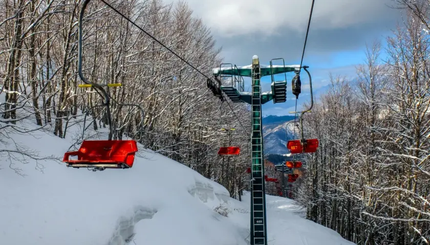 Seggiovie rosse e innevate salgono verso il cielo azzurro, attraversando un bosco spoglio. Un paesaggio invernale che ricorda il **centro** di **Abetone**.