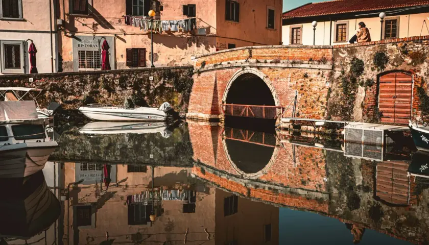 Un ponte in mattoni rossi attraversa un canale nel quartiere Venezia di Livorno. Barche ormeggiate riflettono sulle acque calme, mentre panni stesi aggiungono un tocco di vita quotidiana.