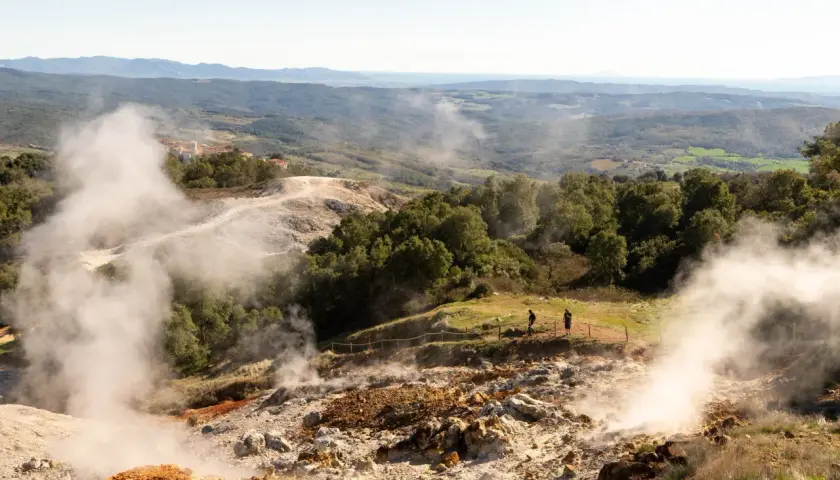 Vapori emergono dal terreno in un paesaggio collinare, con un villaggio in lontananza. Un geyser in Italia che incanta i visitatori.