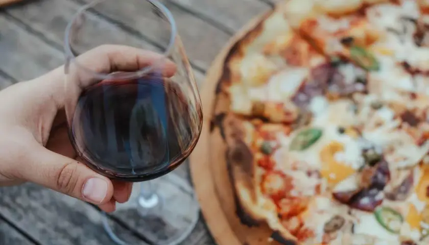 Un bicchiere di vino rosso in mano, accanto a una gustosa pizza. Un abbinamento perfetto per una serata all'aperto.