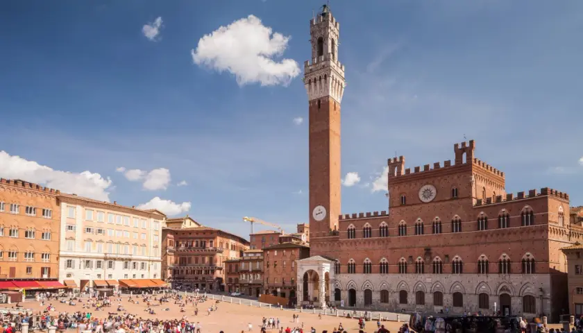 La Piazza del Campo di Siena, con la Torre del Mangia svettante, brulica di gente sotto un cielo azzurro.