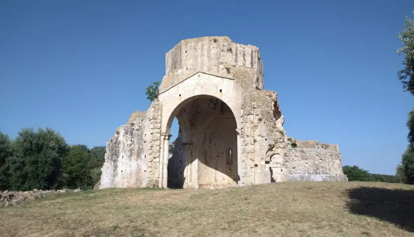 Rovine dell'abbazia di San Bruzio sotto un cielo azzurro, con alberi sullo sfondo.