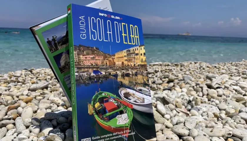 Guida turistica "Isola d'Elba" su una spiaggia di ciottoli, con il mare azzurro sullo sfondo. Perfetta per pianificare la tua vacanza.