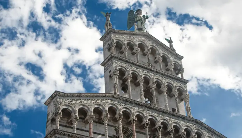 La facciata riccamente decorata della chiesa San Michele a Lucca, con un angelo in cima, si staglia contro un cielo azzurro e nuvoloso.