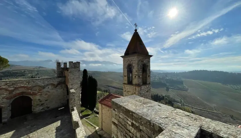 Torre campanaria e mura antiche del castello Frescobaldi, con vigneti e colline toscane sotto un cielo azzurro e soleggiato.