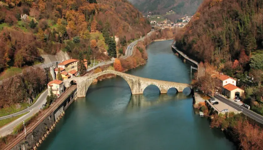 Il Ponte della Maddalena, con i suoi archi irregolari, attraversa un fiume circondato da colline autunnali e case colorate.