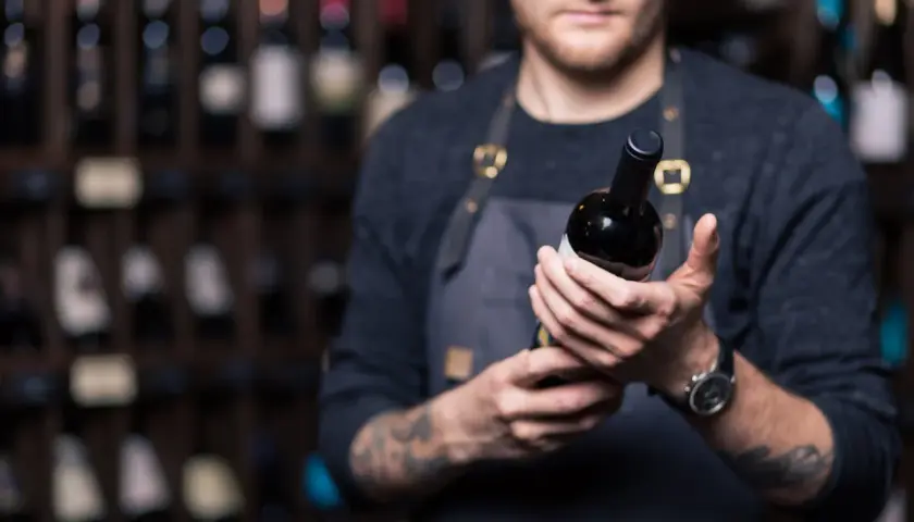 Un sommelier mostra una bottiglia di **vini toscani rossi pregiati**, con scaffali pieni di bottiglie sullo sfondo.