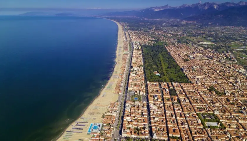 Vista aerea della costa di Massa Carrara, con il mare blu intenso, la lunga spiaggia attrezzata e la città che si estende verso le montagne.