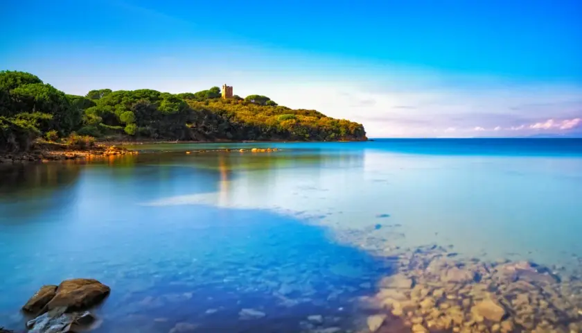 La costa verdeggiante con una torre antica, dove si trova Punta Ala, si specchia nelle acque calme e cristalline del mare.