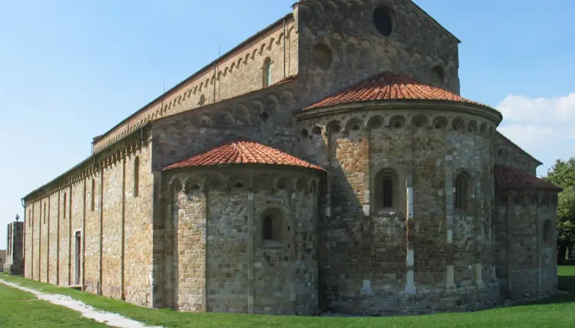 Basilica romanica di San Piero a Grado, con absidi semicircolari e tetto in tegole rosse, su un prato verde sotto un cielo azzurro.