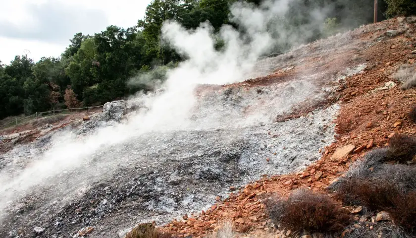 Fumarole di Monterotondo Marittimo emettono vapore su un pendio roccioso e arido, con vegetazione secca e alberi sullo sfondo.