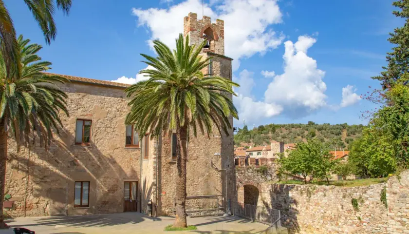 Un antico campanile in pietra a Suvereto, Livorno, svetta tra palme rigogliose e un cielo azzurro con nuvole bianche.