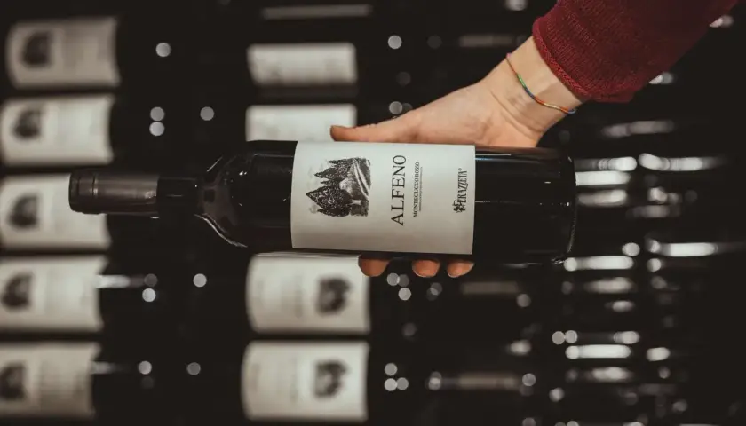 Mano che tiene una bottiglia di vino rosso, etichetta "Alferno Montecucco Rosso", un vino che ricorda il cabernet.