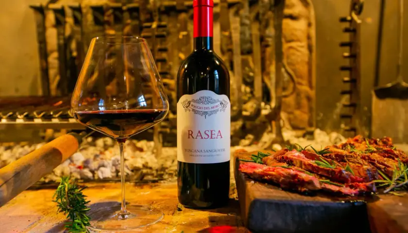 Calice di vino rosso accanto a una bottiglia di Rasea, un vino toscano, con carne grigliata e rosmarino. Un'ode ai super tuscan wines.