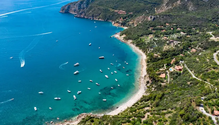 Baia con acque cristalline e numerose barche, incastonata tra la costa verdeggiante e le pendici del monte Argenia.