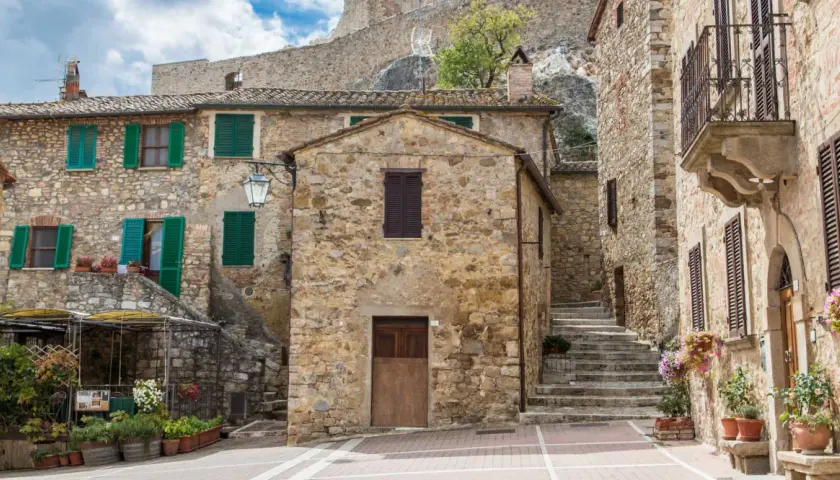 Un vicolo pittoresco a Castel d'Orcia, con case in pietra, persiane verdi e fiori colorati.