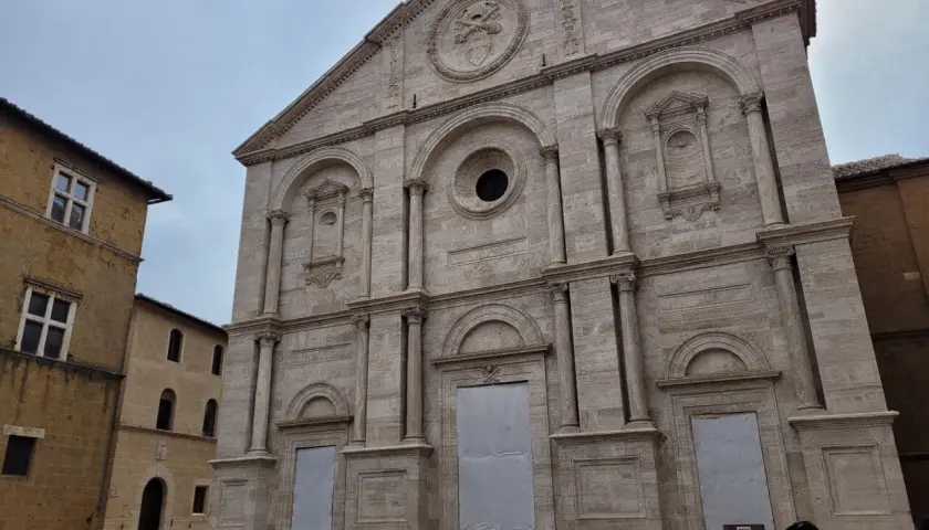 Facciata imponente del Duomo di Pienza, con accanto un edificio in pietra che ricorda il Palazzo Piccolomini.
