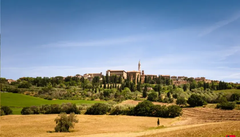 Un borgo medievale domina la Val d'Orcia, circondato da campi dorati e cipressi sotto un cielo azzurro.