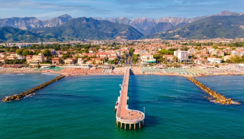 Un molo si estende nel mare turchese di Marina di Massa, con spiagge affollate e montagne sullo sfondo.