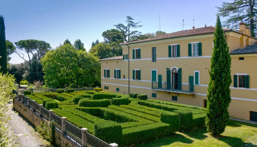 Villa Gracciano della Seta: un giallo edificio con un giardino geometrico di siepi verdi e un cipresso slanciato.