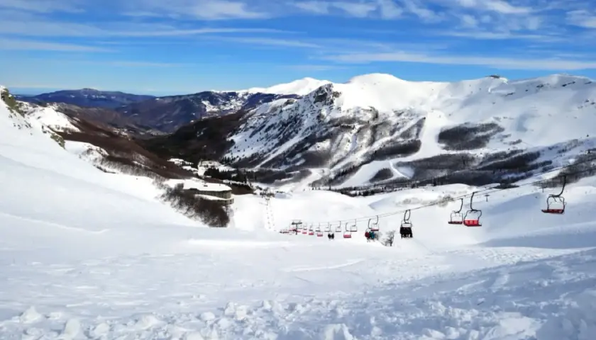 Impianti di risalita rossi trasportano sciatori su piste innevate ad Abetone, con montagne sullo sfondo sotto un cielo azzurro.