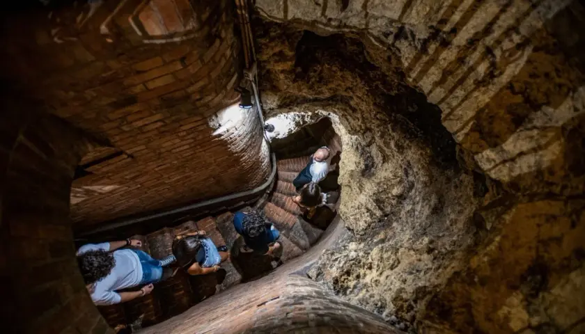 Persone esplorano le antiche cantine storiche di Montepulciano, scendendo una scala a chiocciola in mattoni e tufo.