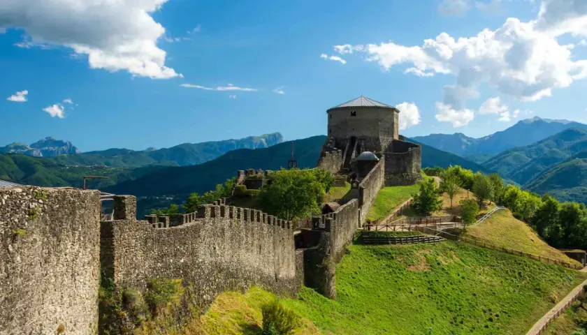 Roccia imponente e mura antiche del **San Romano in Garfagnana** si stagliano su un paesaggio verdeggiante, sotto un cielo azzurro con nuvole bianche.