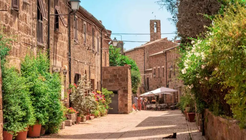 Un gatto nero riposa al sole in un vicolo fiorito della Maremma Grossetana. Case in pietra, lampioni e un campanile completano la scena.